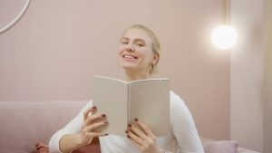A cheerful woman in a cozy setting smiles while holding and reading a notebook indoors.