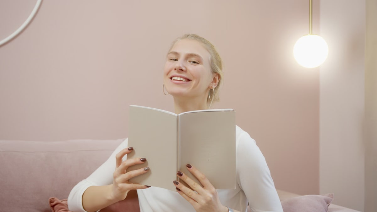 A cheerful woman in a cozy setting smiles while holding and reading a notebook indoors.